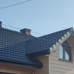 Nowy dach z czarnej blachy trapezowej na domu jednorodzinnym w Niepołomicach, widoczny komin z cegły oraz ozdobny wiatrowskaz na szczycie dachu.