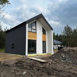 AGMAR Marcin Karpinski - Nowoczesny dom jednorodzinny z czarno-białą elewacją i drewnianymi akcentami, widoczny w trakcie wykończenia, z ziemią i stertą gruzu wokół.