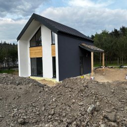 AGMAR Marcin Karpinski - Nowoczesny dom w trakcie wykańczania elewacji: biała i grafitowa fasada, drewniane panele, widoczna sterta ziemi przed budynkiem, teren budowy.