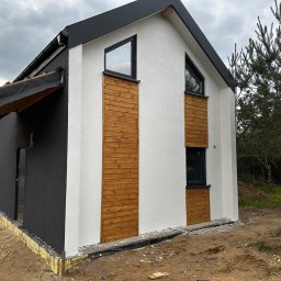 AGMAR Marcin Karpinski - Nowoczesny dom jednorodzinny z białą elewacją, drewnianymi panelami pionowymi wokół okien i czarną elewacją boczną z zadaszeniem w trakcie budowy, widoczne elementy izolacji przy gruncie.