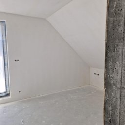 AGMAR Marcin Karpinski - Wnętrze w trakcie wykańczania: ściany pokryte świeżą, jasną gładzią, widoczne puszki elektryczne, surowy betonowy filar w rogu. Na podłodze widoczne ślady po pracach budowlanych i butelka z wodą.