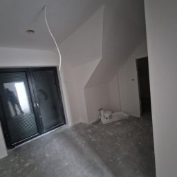 AGMAR Marcin Karpinski - Surowe wnętrze poddasza z oknem balkonowym, ścianami po gładziowaniu i niedokończoną instalacją elektryczną, widoczne ślady po pracach remontowych na podłodze.