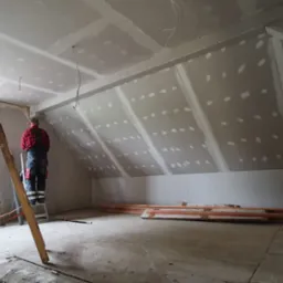 Prace wykończeniowe na poddaszu: montaż płyt gipsowo-kartonowych na skośnej ścianie, widoczny pracownik na drabinie w czerwonej koszuli w kratę, szpachlowane łączenia płyt, przygotowane profile...