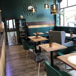 AGMAR Marcin Karpinski - Wnętrze kawiarni z zieloną ścianą, drewnianymi stolikami, welurowymi krzesłami i dekoracyjnym lustrem nad zieloną kanapą. Na ścianie wisi oprawiona kolekcja pudełek od zapałek.