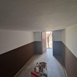 AGMAR Marcin Karpinski - Wnętrze pomieszczenia z częściowo wykończonymi ścianami, gdzie dolna część jest w kolorze brązowym, a górna biała. Na podłodze leżą narzędzia malarskie, a w otwartych drzwiach stoi osoba...