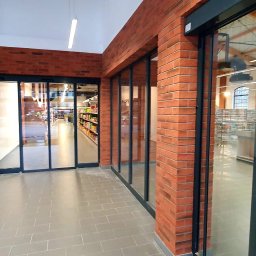 AGMAR Marcin Karpinski - Nowoczesne wejście do sklepu z automatycznymi drzwiami przesuwnymi, wykończone czerwoną cegłą i szarymi płytkami podłogowymi.