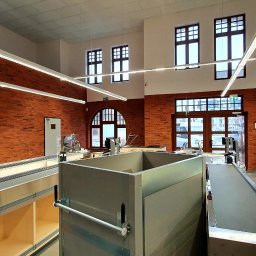 AGMAR Marcin Karpinski - Wnętrze budynku z wyeksponowaną ścianą z czerwonej cegły, widoczne okna z ciemnymi ramami, jasne, podłużne lampy sufitowe oraz elementy wyposażenia sklepowego, takie jak stanowiska kasowe...