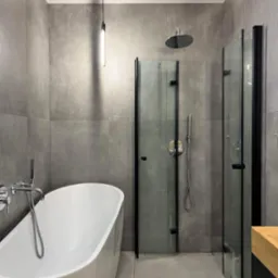 Nowoczesna łazienka z wolnostojącą białą wanną, szarymi płytkami imitującymi beton, kabiną prysznicową z czarnymi ramami i minimalistyczną lampą wiszącą.