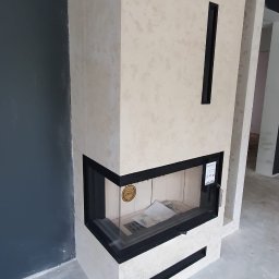 AGMAR Marcin Karpinski - Narożny kominek obudowany płytami imitującymi trawertyn z czarnymi detalami, widoczny na tle szarej ściany i surowej posadzki.