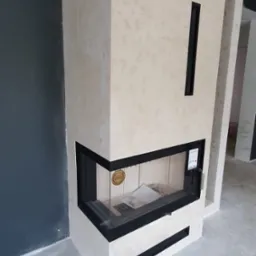 Narożny kominek obudowany płytami imitującymi trawertyn z czarnymi detalami, widoczny na tle szarej ściany i surowej posadzki.