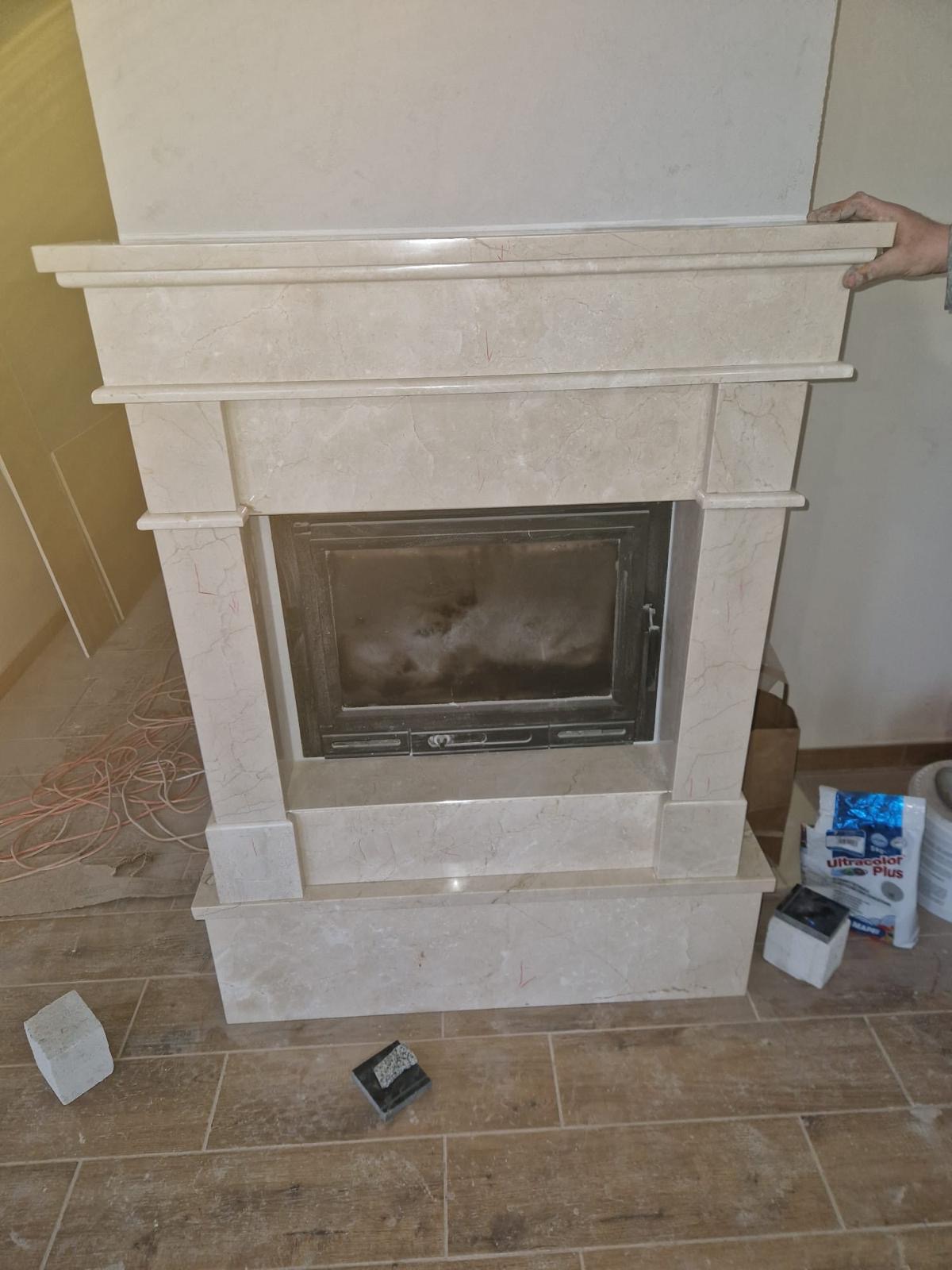 Marmurowa obudowa kominka z czarnym wkładem kominkowym, widoczne narzędzia i materiały budowlane na podłodze wyłożonej płytkami imitującymi drewno, ręka osoby montującej widoczna w prawym górnym rogu.