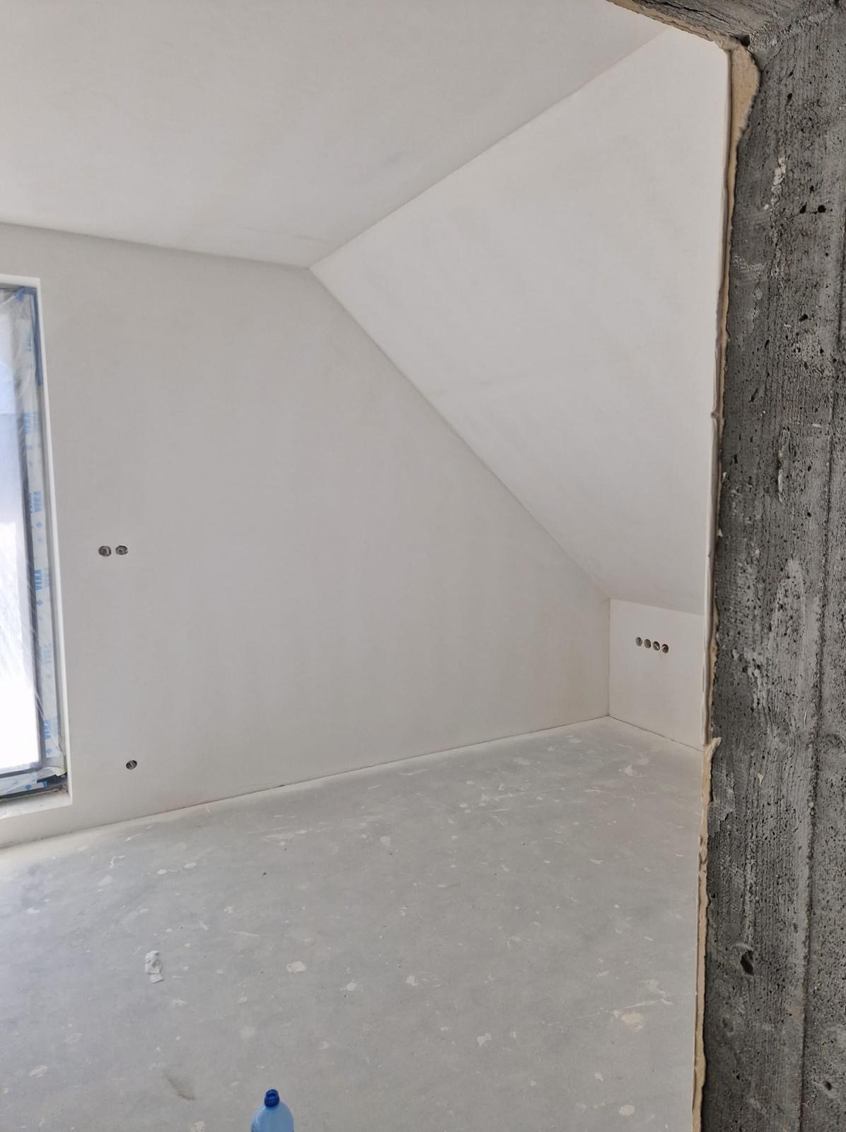 Wnętrze w trakcie wykańczania: ściany pokryte świeżą, jasną gładzią, widoczne puszki elektryczne, surowy betonowy filar w rogu. Na podłodze widoczne ślady po pracach budowlanych i butelka z wodą.