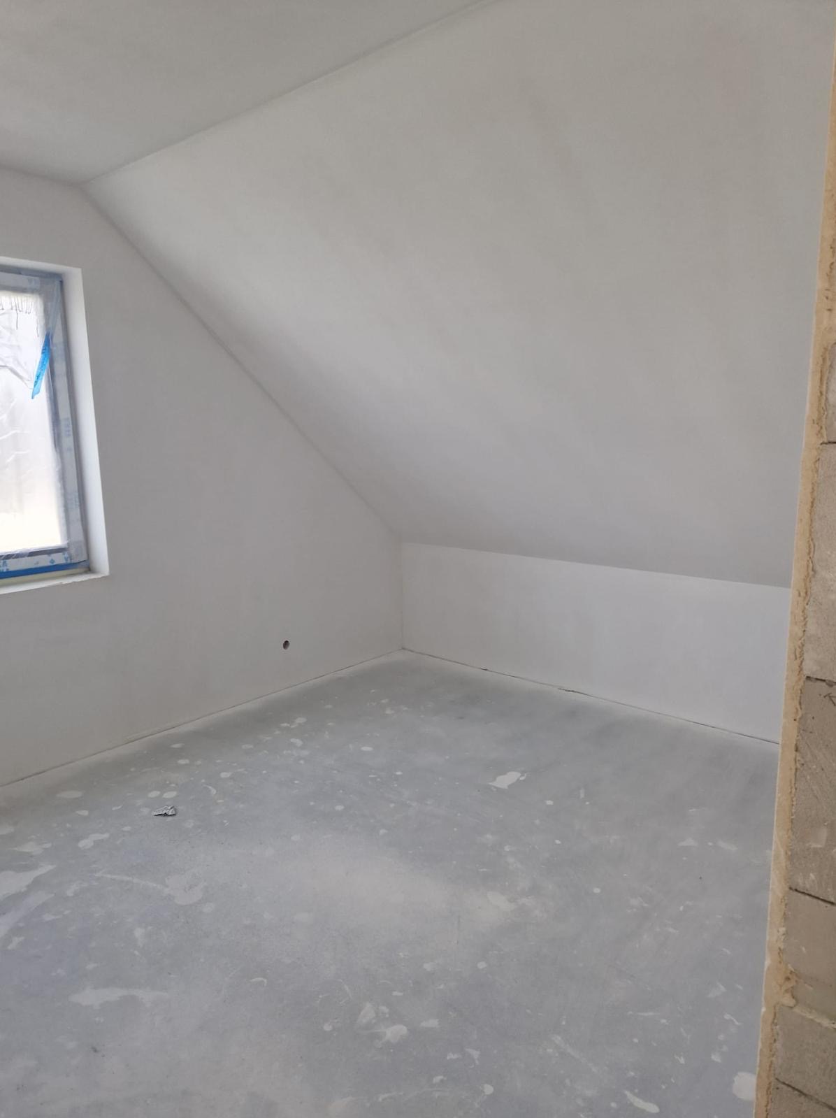 Poddasze po nałożeniu gładzi, widoczne ślady po pracach na podłodze, okno zabezpieczone folią malarską, ściany i sufit pomalowane na biało.
