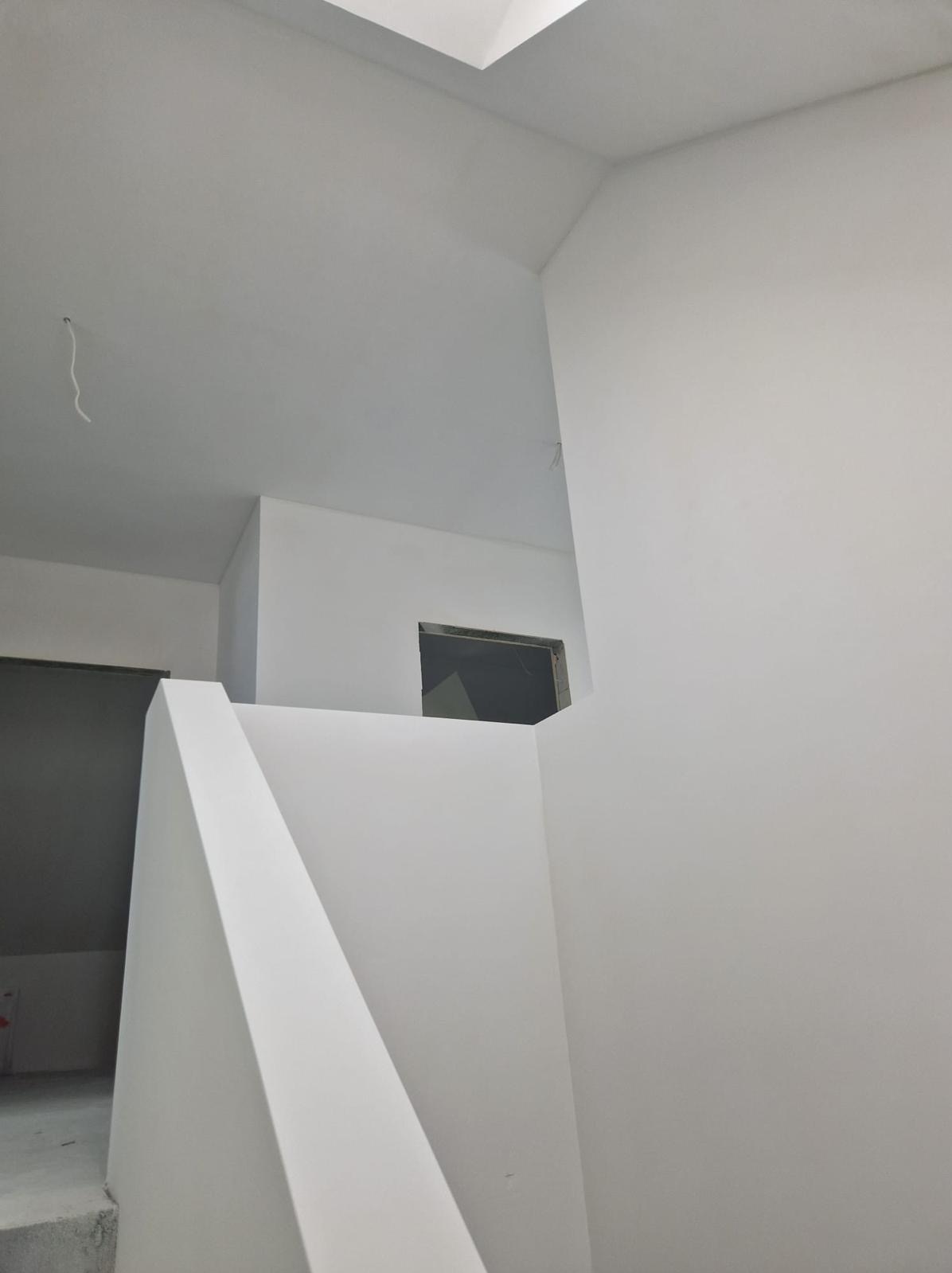 Wnętrze klatki schodowej po malowaniu na biało, widoczne gładkie ściany i narożniki, z niedokończonym otworem okiennym.
