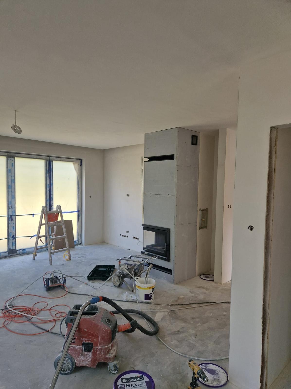 Wnętrze w trakcie remontu: ściany po nałożeniu gładzi, zabudowa kominka z płyt gipsowo-kartonowych, drabina oparta o okno, sprzęt malarski, odkurzacz przemysłowy i wiaderko z farbą.