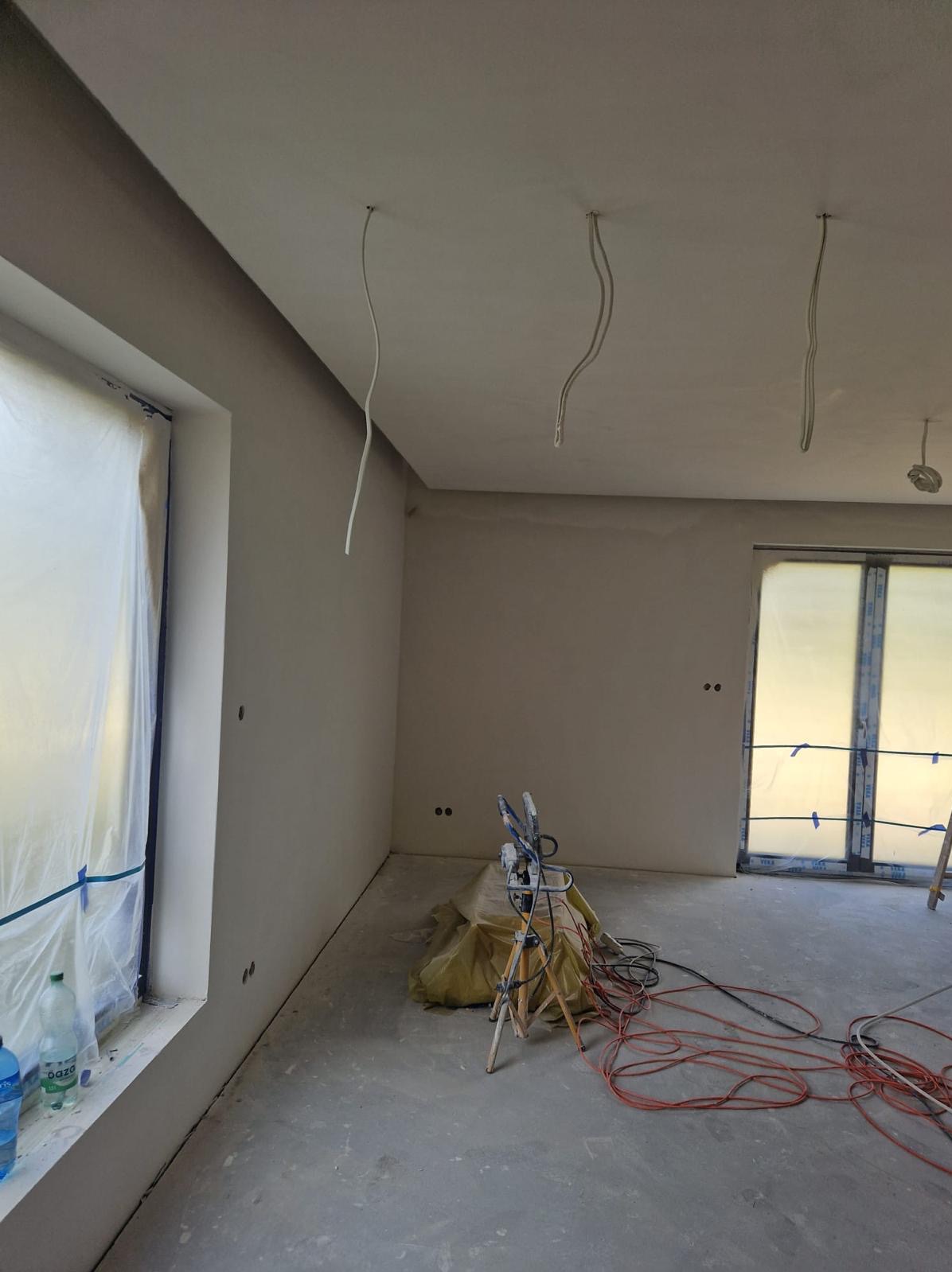 Pokój w trakcie prac wykończeniowych, ściany po nałożeniu gładzi, okno zabezpieczone folią, widoczny agregat malarski i przewody na podłodze, przygotowanie do malowania.