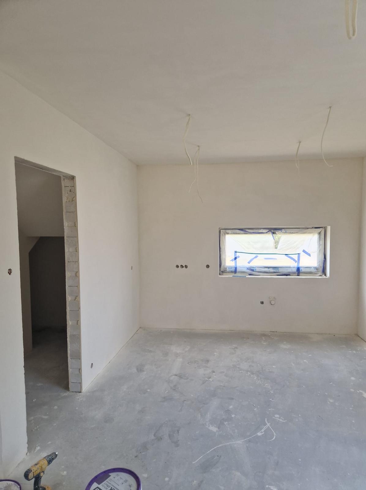 Wnętrze w trakcie remontu: gładkie, białe ściany, surowy beton na podłodze, okno zabezpieczone folią, wystające kable ze ściany i sufitu, puszki z farbą i wiertarka na podłodze w rogu.