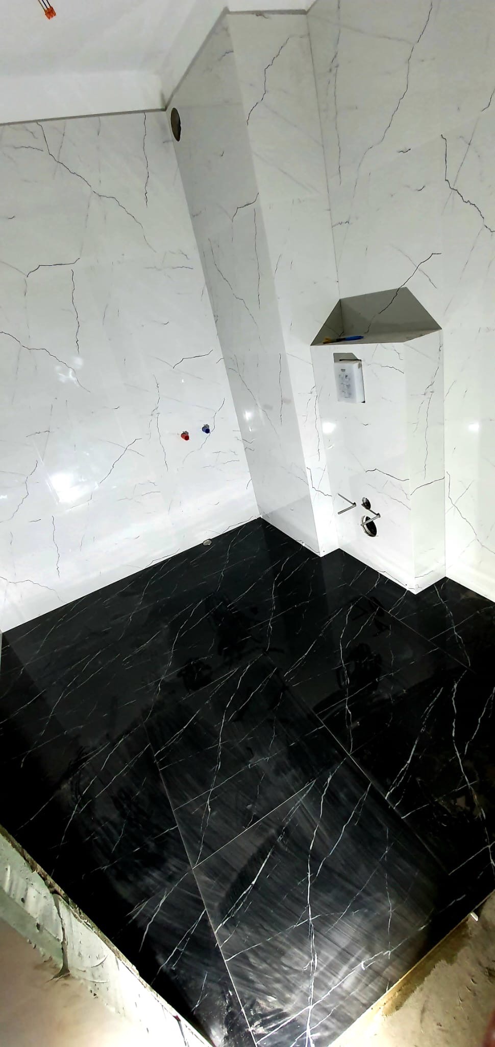 Wykończona łazienka z czarną podłogą z płytek imitujących marmur oraz białymi ścianami z płytek z szarymi żyłami, widoczne przyłącza wodne i wnęka w ścianie.