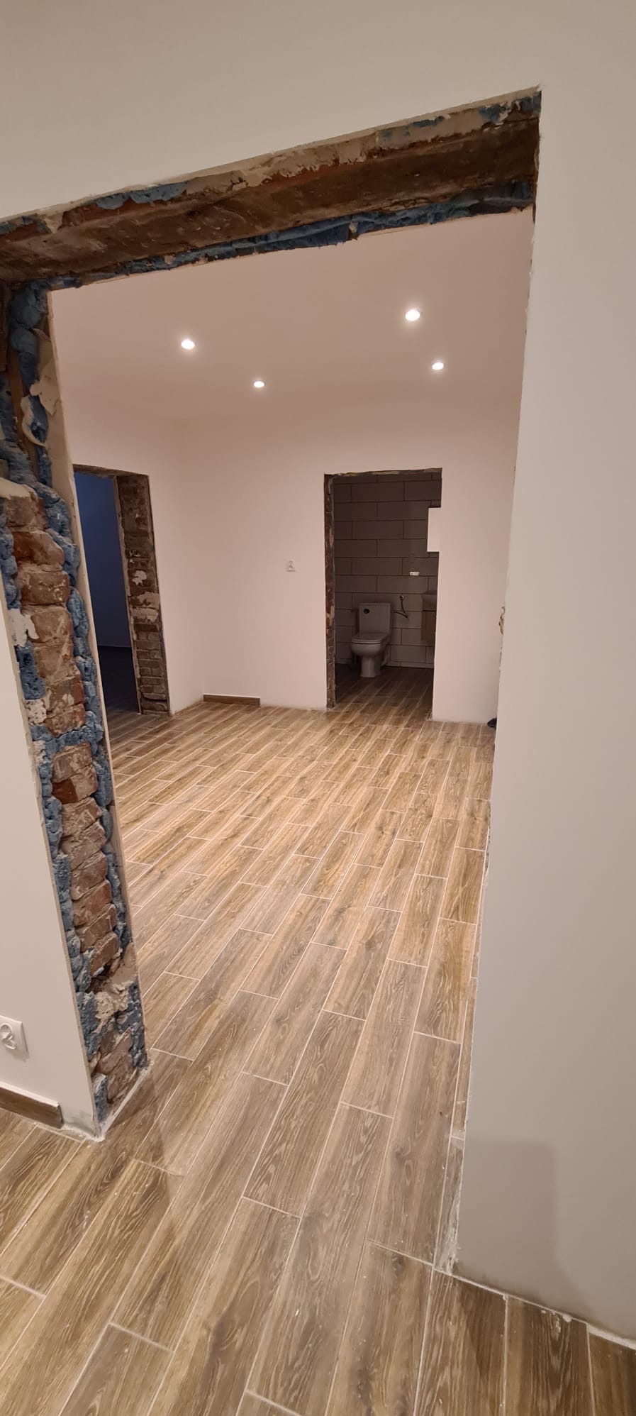 Wnętrze w trakcie remontu z ułożoną podłogą z płytek imitujących drewno, widoczne surowe otwory drzwiowe z odsłoniętą cegłą i fragment łazienki z toaletą w głębi.