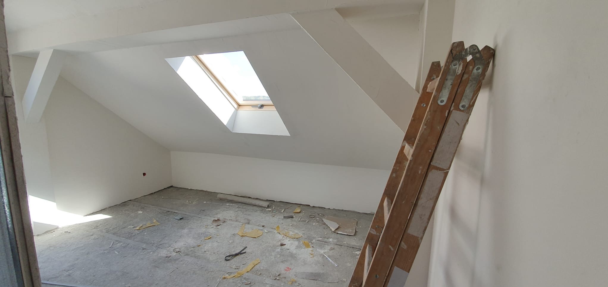 Poddasze po świeżo wykonanych pracach wykończeniowych, widoczne gładkie, białe ściany, okno dachowe i drewniana drabina oparta o ścianę. Na podłodze resztki materiałów budowlanych.