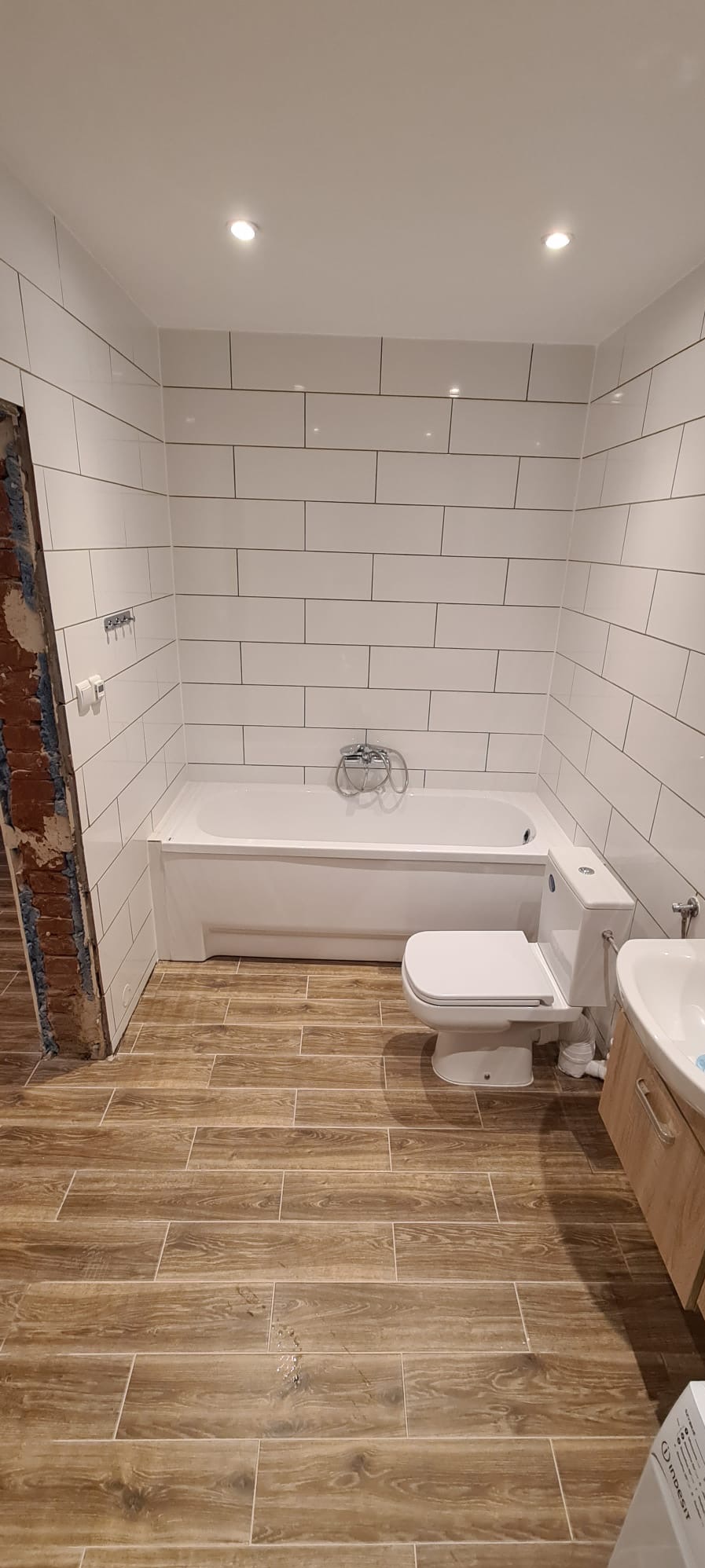 Wykończona łazienka z białymi, prostokątnymi płytkami na ścianach, drewnopodobnymi płytkami na podłodze, białą wanną, toaletą i umywalką z szafką w kolorze jasnego drewna. Widoczny fragment ściany...