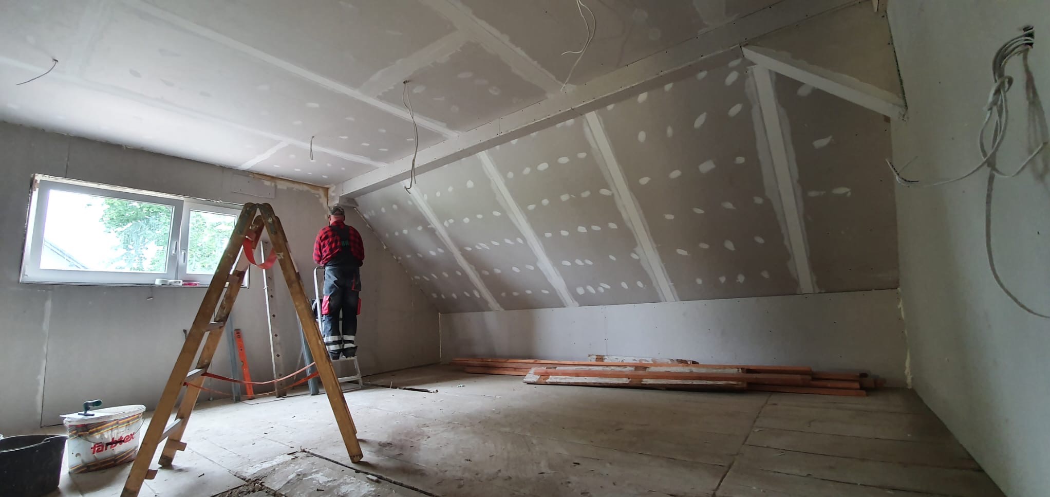 Prace wykończeniowe na poddaszu: montaż płyt gipsowo-kartonowych na skośnej ścianie, widoczny pracownik na drabinie w czerwonej koszuli w kratę, szpachlowane łączenia płyt, przygotowane profile...