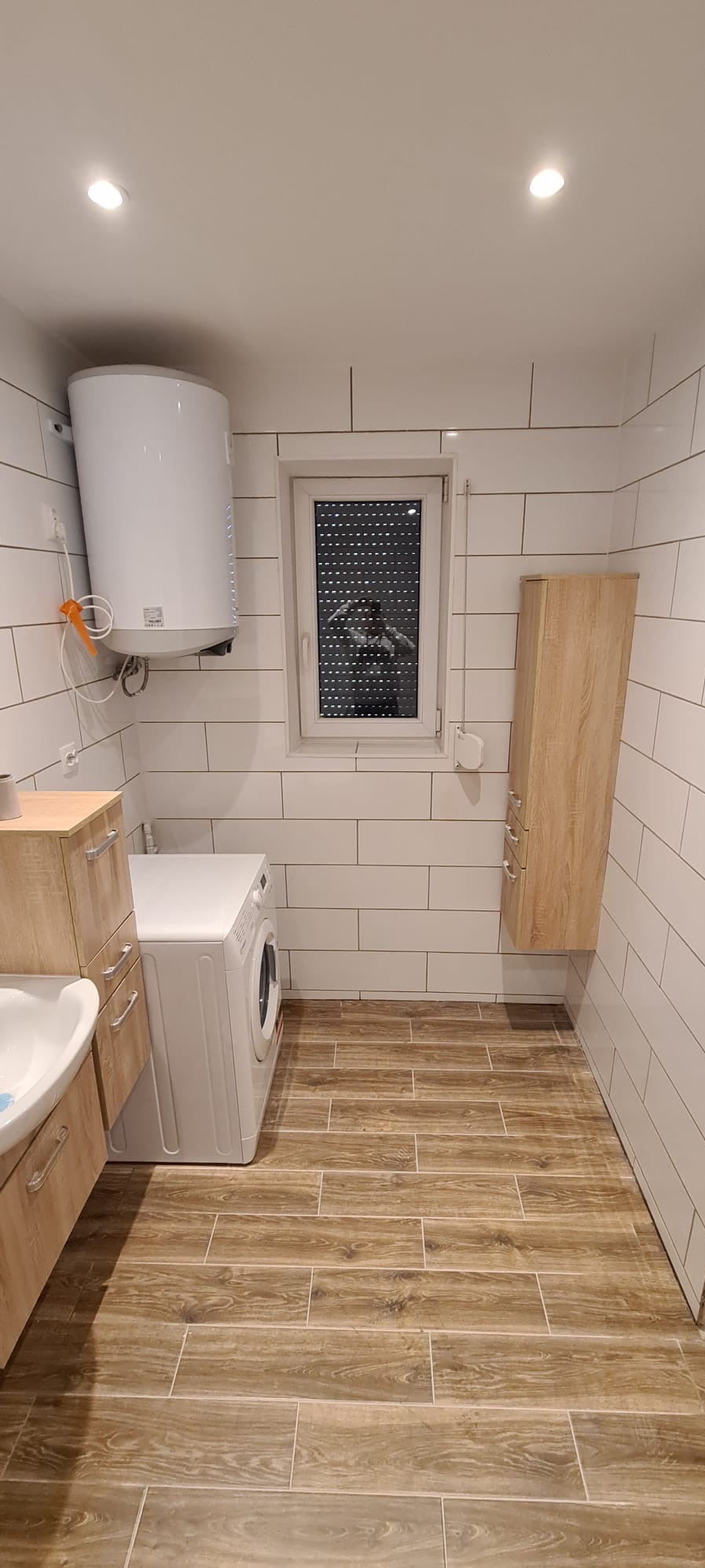 Nowoczesna łazienka z białymi płytkami, drewnopodobną podłogą, pralką, bojlerem i szafką wiszącą; w oknie widoczne odbicie fotografa.