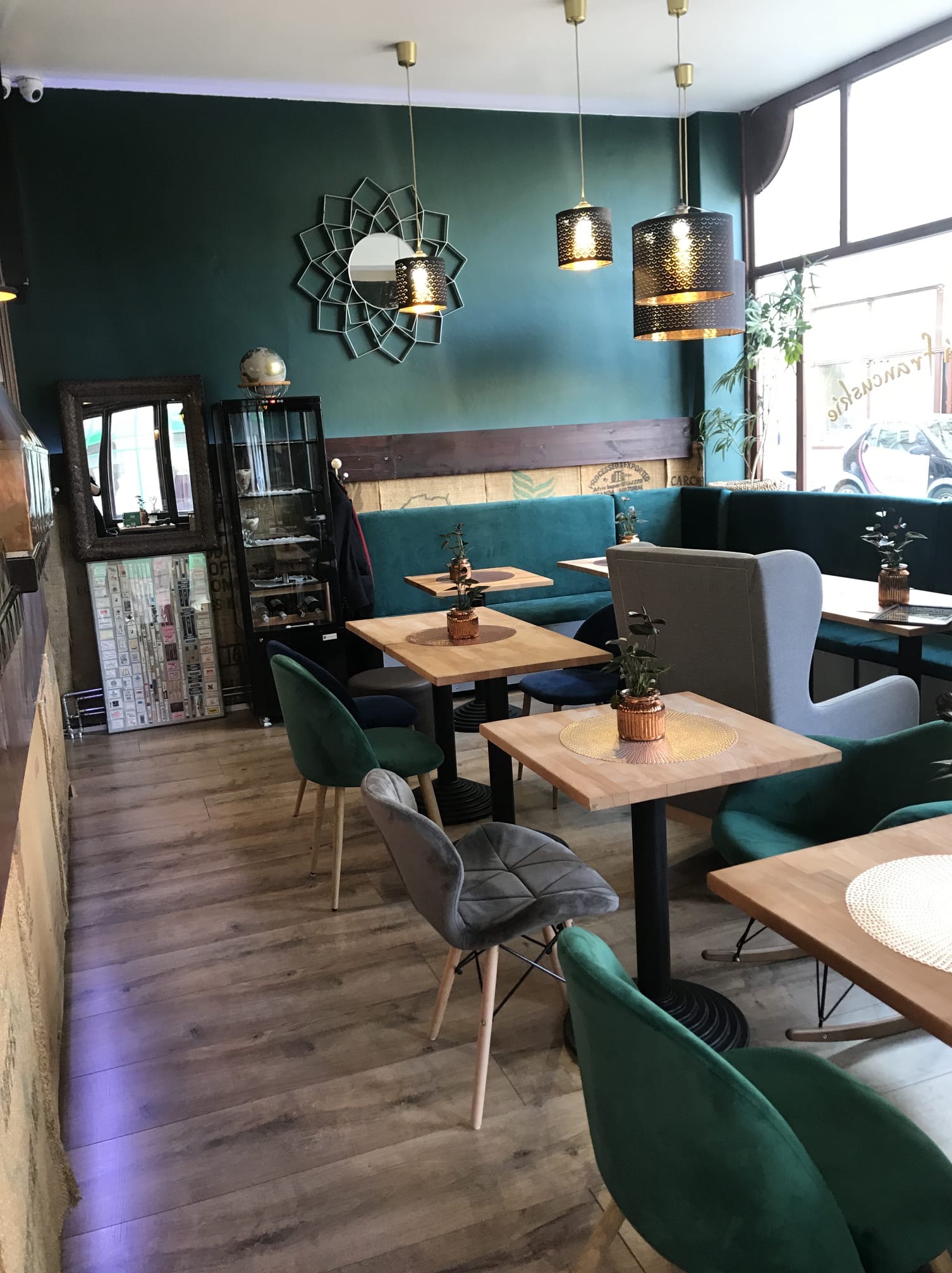 Wnętrze kawiarni z zieloną ścianą, drewnianymi stolikami, welurowymi krzesłami i dekoracyjnym lustrem nad zieloną kanapą. Na ścianie wisi oprawiona kolekcja pudełek od zapałek.