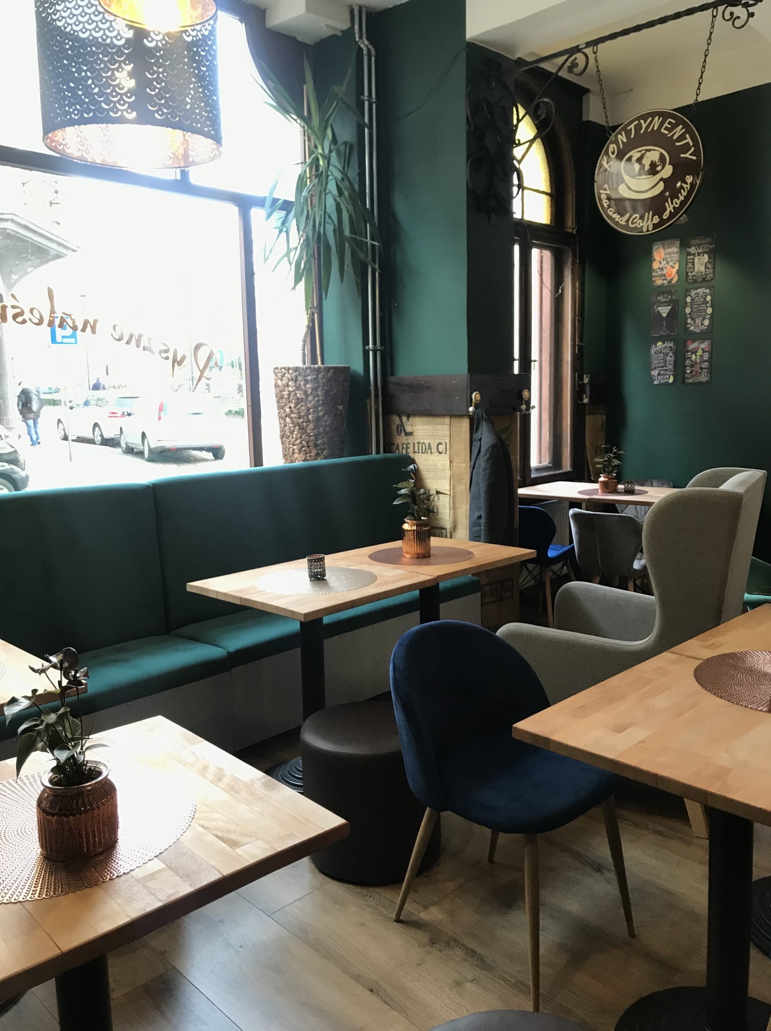 Wnętrze kawiarni w Lisewo z zielonymi ścianami, drewnianymi stolikami, tapicerowanymi krzesłami i kanapą oraz roślinami doniczkowymi.
