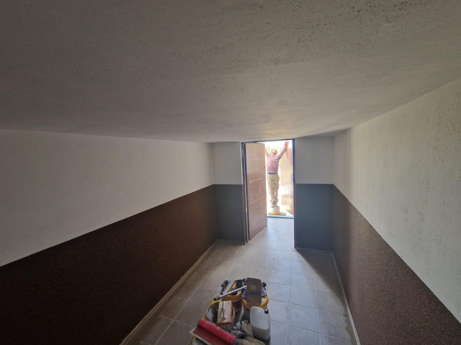 Wnętrze pomieszczenia z częściowo wykończonymi ścianami, gdzie dolna część jest w kolorze brązowym, a górna biała. Na podłodze leżą narzędzia malarskie, a w otwartych drzwiach stoi osoba...