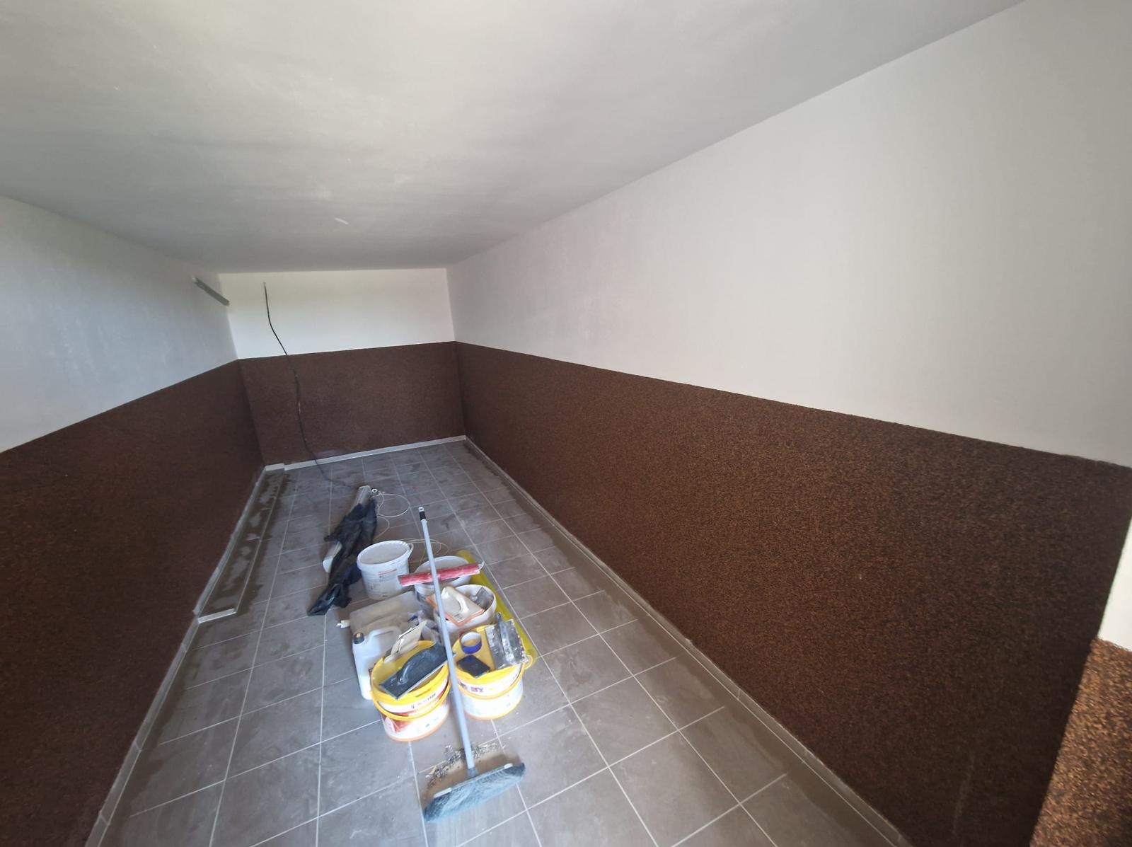 Wnętrze pomieszczenia po częściowym wykończeniu: dolna część ścian pokryta brązową strukturą, górna część pomalowana na biało, widoczne narzędzia i wiaderka z farbą na podłodze z płytek.