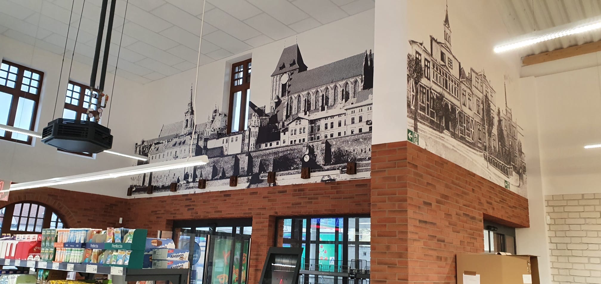 Wnętrze sklepu z dekoracyjną fototapetą przedstawiającą czarno-biały krajobraz miejski, z elementami muru z cegły i widocznymi półkami z produktami.