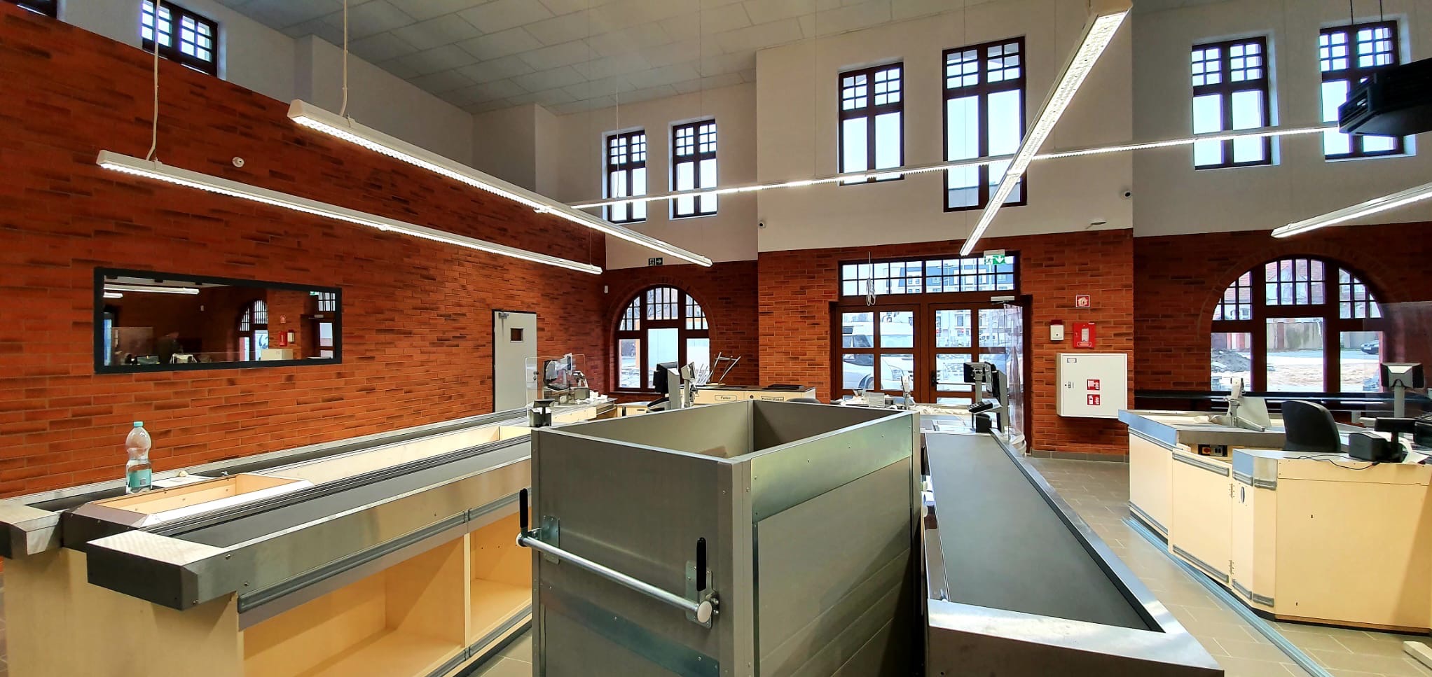 Wnętrze budynku z wyeksponowaną ścianą z czerwonej cegły, widoczne okna z ciemnymi ramami, jasne, podłużne lampy sufitowe oraz elementy wyposażenia sklepowego, takie jak stanowiska kasowe...