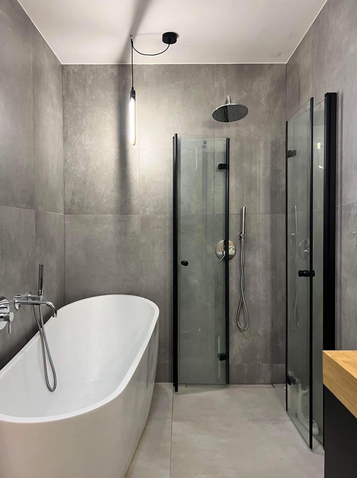 Nowoczesna łazienka z wolnostojącą białą wanną, szarymi płytkami imitującymi beton, kabiną prysznicową z czarnymi ramami i minimalistyczną lampą wiszącą.