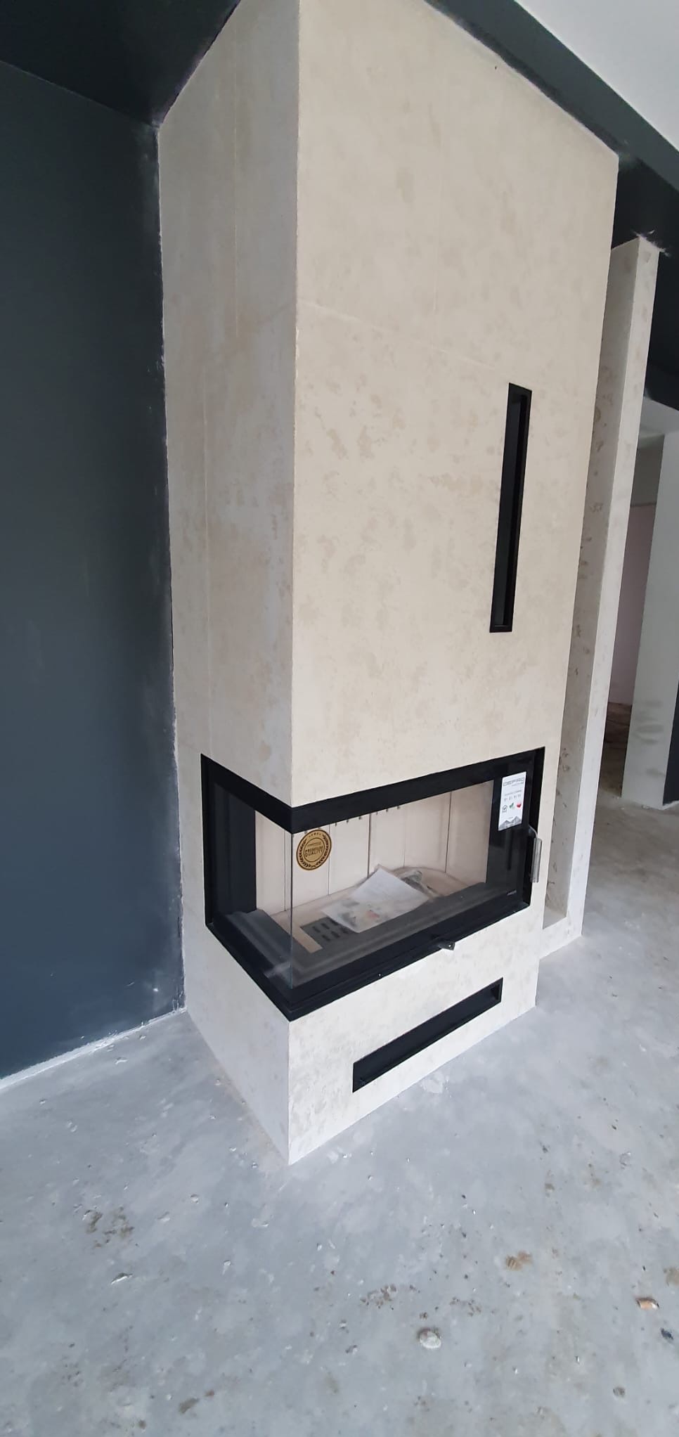 Narożny kominek obudowany płytami imitującymi trawertyn z czarnymi detalami, widoczny na tle szarej ściany i surowej posadzki.