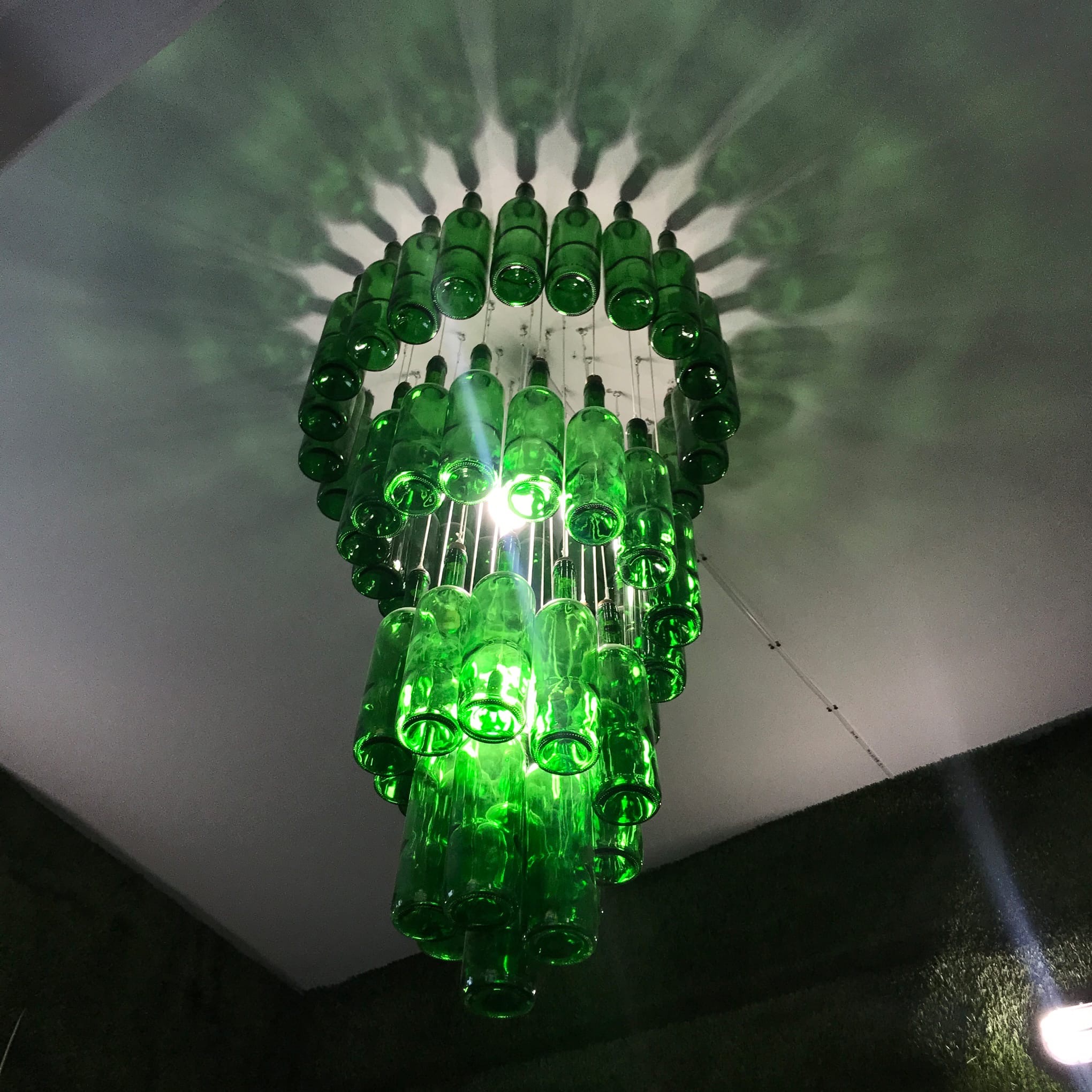 Nietypowy żyrandol wykonany z zielonych butelek po napojach, zawieszony pod sufitem, oświetlony od wewnątrz, rzucający cienie na sufit.