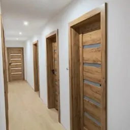 Korytarz po remoncie z zamontowanymi drzwiami z drewnianą okleiną i przeszkleniami, jasne panele podłogowe, białe ściany i sufit z oświetleniem punktowym.