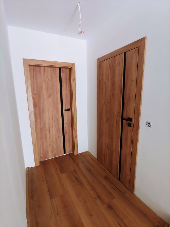 Dwa zamontowane drzwi wewnętrzne z okleiną drewnopodobną i czarnymi akcentami, widoczne na tle białych ścian i podłogi z paneli w odcieniach brązu. Na suficie widoczny wystający kabel.