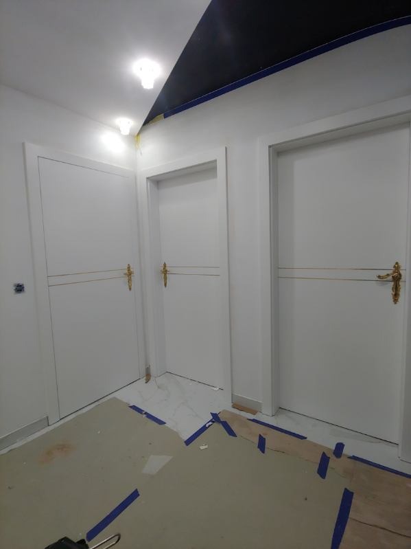 Trzy białe, panelowe drzwi z złotymi klamkami w trakcie montażu, zabezpieczone niebieską taśmą malarską na podłodze.