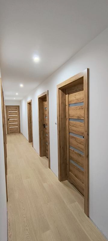 Korytarz po remoncie z zamontowanymi drzwiami z drewnianą okleiną i przeszkleniami, jasne panele podłogowe, białe ściany i sufit z oświetleniem punktowym.