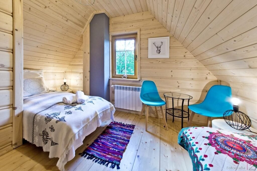 Drewniany pokój na poddaszu z dwoma turkusowymi krzesłami, łóżkiem z narzutą w etniczne wzory, oknem i obrazkiem jelenia na ścianie.