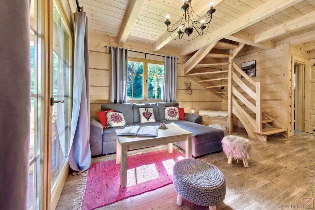 Przytulny salon w drewnianym domku z narożną sofą, spiralnymi schodami i różowym dywanem, oświetlony naturalnym światłem wpadającym przez okno.