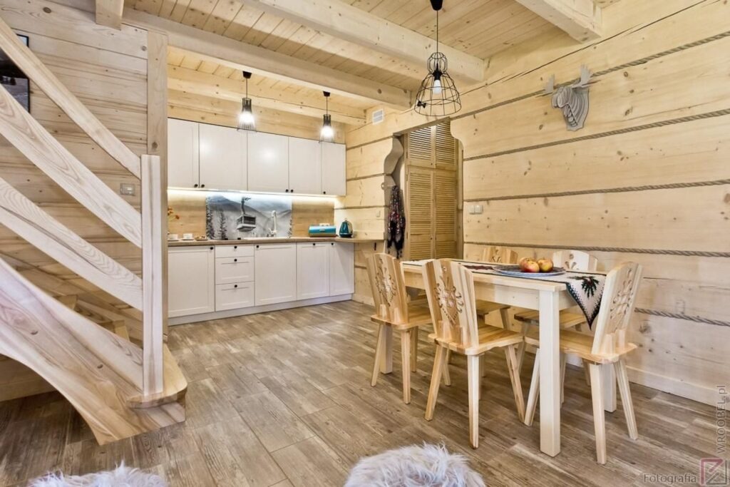 Jasne, drewniane wnętrze w stylu góralskim: schody, kuchnia z białymi szafkami, stół z krzesłami z dekoracyjnymi wycięciami, drewniana dekoracja w kształcie głowy jelenia na ścianie.