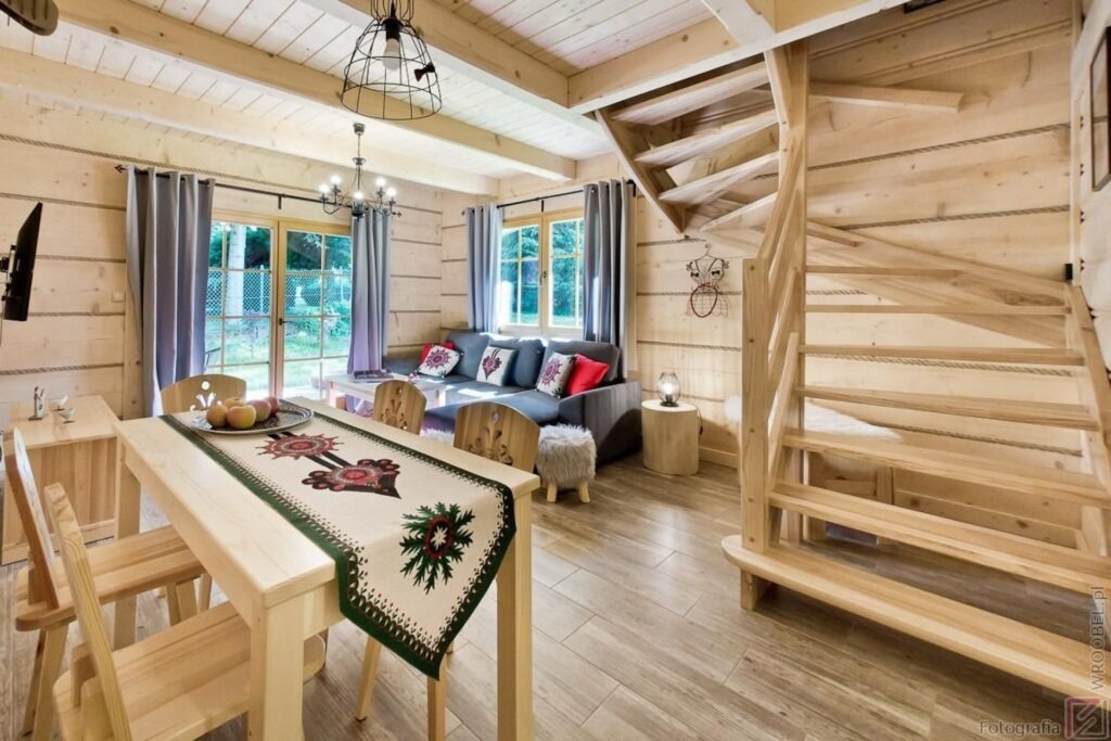 Jasne, drewniane wnętrze domu z widocznymi schodami, stołem z dekoracyjnym bieżnikiem i szarą sofą z kolorowymi poduszkami.