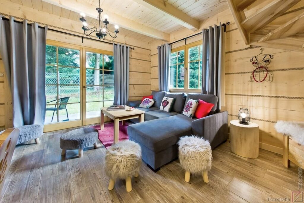 Przytulne wnętrze drewnianego domku z szarym narożnikiem, puszystymi stołkami, drewnianymi ścianami i oknami z widokiem na zieleń.