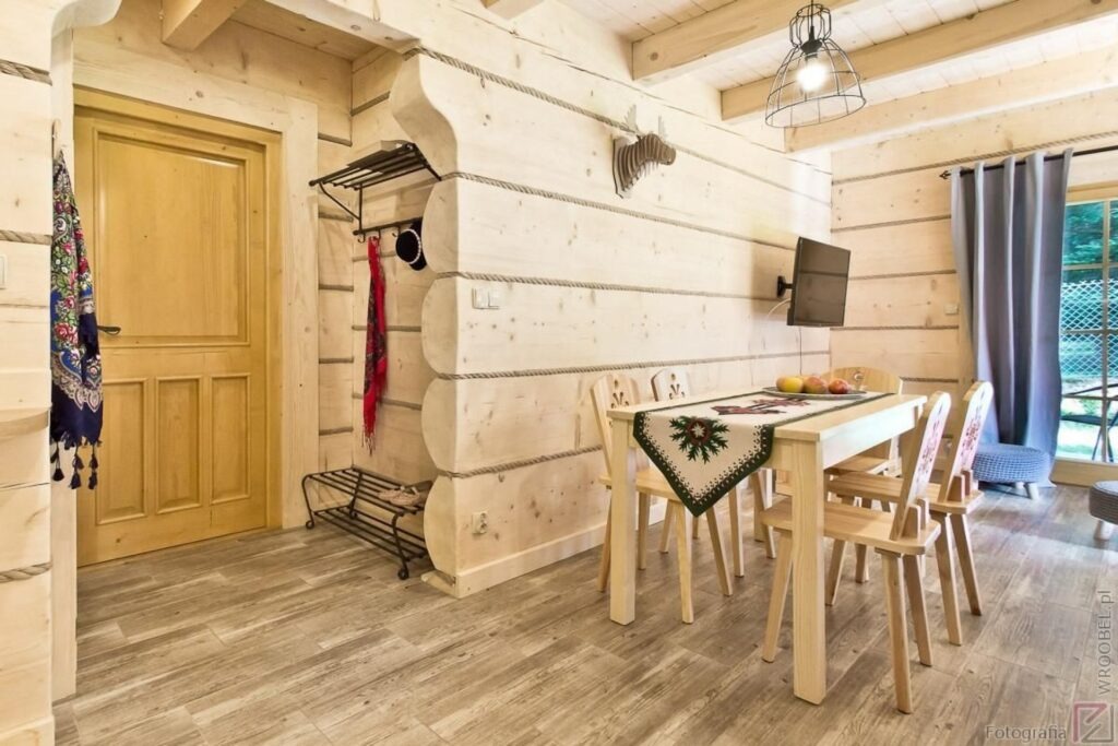 Wnętrze drewnianego domku z jasnymi ścianami, drewnianym stołem z dekoracyjnym obrusem, krzesłami i telewizorem na ścianie, w tle okno z zasłoną.