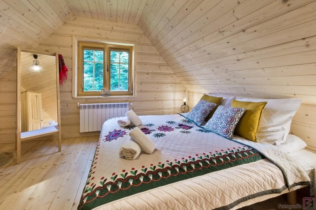 Drewniana sypialnia na poddaszu z oknem, grzejnikiem, lustrem i łóżkiem z wzorzystą narzutą oraz ręcznikami.