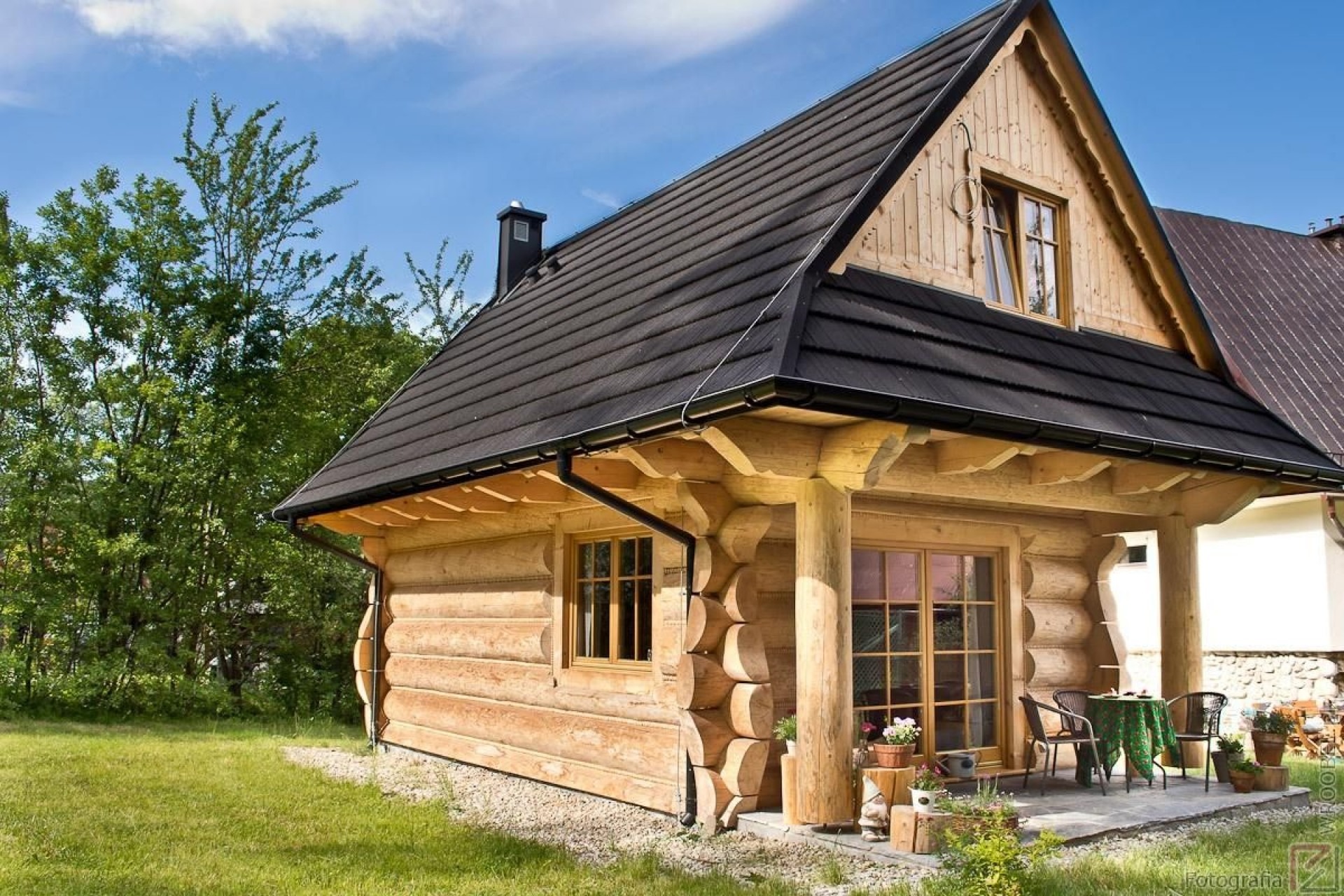 Drewniany domek letniskowy z ciemnym dachem i małym tarasem, otoczony zielenią trawnika i drzew.