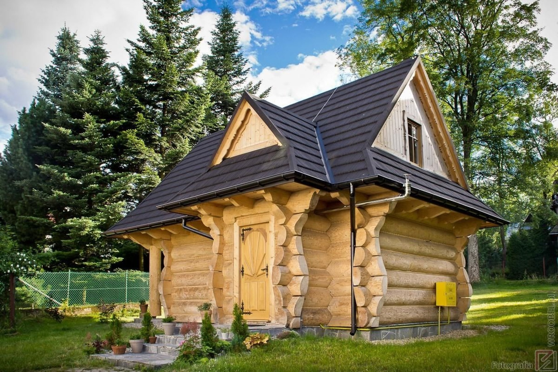 Mały, drewniany domek letniskowy z ciemnym dachem i jasnymi drzwiami, otoczony zielenią drzew i trawnikiem, z widoczną skrzynką gazową przy ścianie.