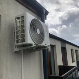 Biała jednostka zewnętrzna klimatyzacji Rotenso Ukura zamontowana na ścianie budynku mieszkalnego z widocznym fragmentem balkonu i pochmurnym niebem w tle.